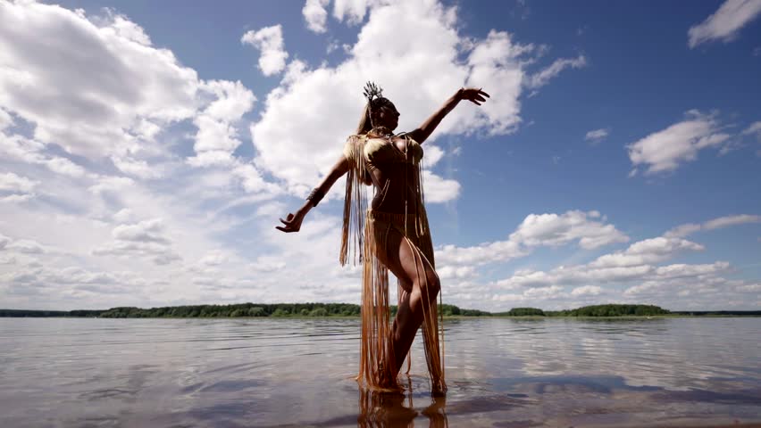Against the backdrop of a picturesque lake and a blue sky with clouds, a woman in an exotic costume with a crown dances in the water, her movements are graceful and light.