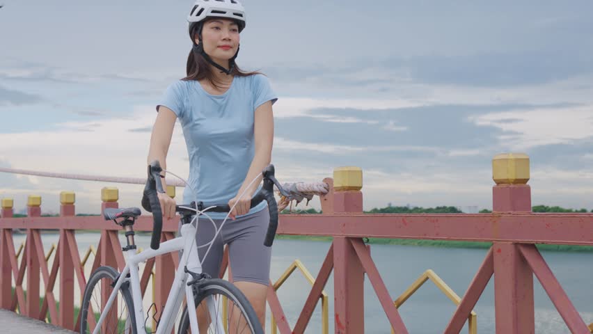 Asian young woman pushing bicycle up the hill on the bridge in garden. Attractive beautiful sportswoman in sportswear feel happy and relax, biking exercise workout for health activity in public park.