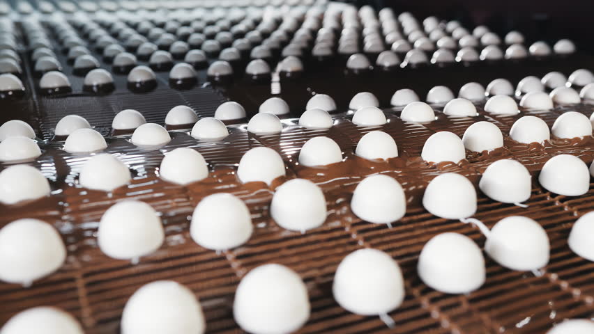 White candy pieces moving along a conveyor and getting coated with chocolate in a modern confectionery production line inside an automated factory environment.