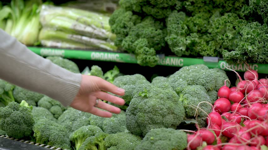 A woman in a beige sweater selecting fresh broccoli from a display in the produce section of a grocery store, with other vegetables such as radishes and kale nearby.