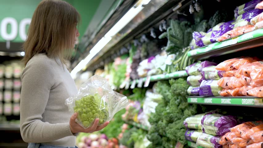 A woman in a beige sweater and blue jeans holding a package of lettuce in the produce section of a grocery store, surrounded by various fresh vegetables and packaged items.