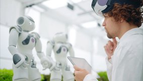 Laboratory technician in a white coat wearing virtual reality headset analysing graphs on a tablet while interacting with two humanoid robots in a greenhouse farm - Powered by Shutterstock - Get 15% off with code: PIKWIZARD15