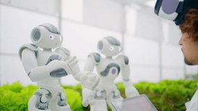 Laboratory technician in a white coat wearing virtual reality headset analysing graphs on a tablet while interacting with two humanoid robots in a greenhouse farm - Powered by Shutterstock - Get 15% off with code: PIKWIZARD15