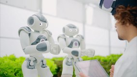 Laboratory technician in a white coat wearing virtual reality headset analysing graphs on a tablet while interacting with two humanoid robots in a greenhouse farm - Powered by Shutterstock - Get 15% off with code: PIKWIZARD15