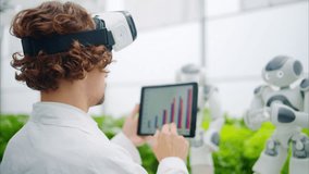 Laboratory technician in a white coat wearing virtual reality headset analysing graphs on a tablet while interacting with two humanoid robots in a greenhouse farm - Powered by Shutterstock - Get 15% off with code: PIKWIZARD15