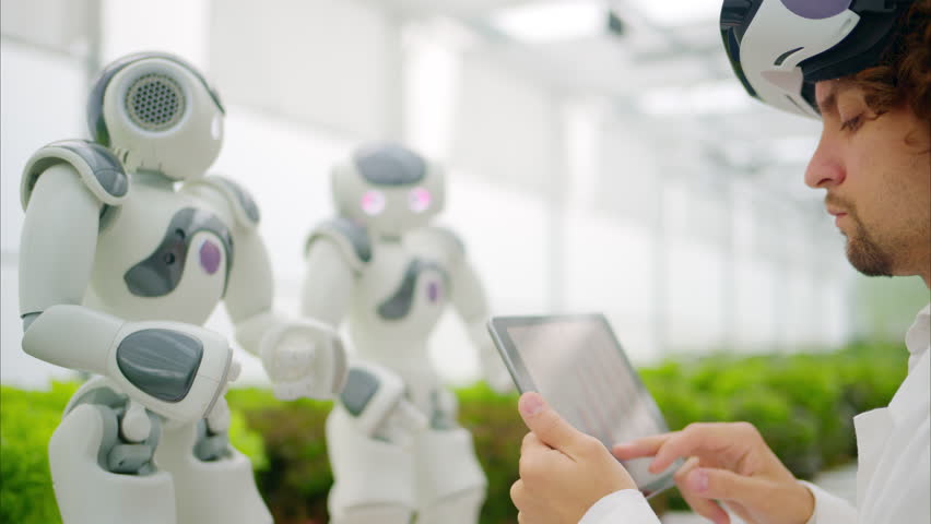 Laboratory technician in a white coat wearing virtual reality headset analysing graphs on a tablet while interacting with two humanoid robots in a greenhouse farm - Powered by Shutterstock - Get 15% off with code: PIKWIZARD15