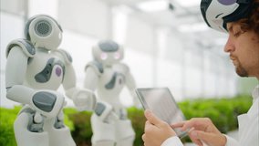 Laboratory technician in a white coat wearing virtual reality headset analysing graphs on a tablet while interacting with two humanoid robots in a greenhouse farm - Powered by Shutterstock - Get 15% off with code: PIKWIZARD15