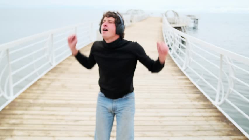 Cute young guy party-goer enjoying music in headphones, having fun dancing standing on the shore of the lake in happy solitude with his arms outstretched, celebrating acceptance to university.