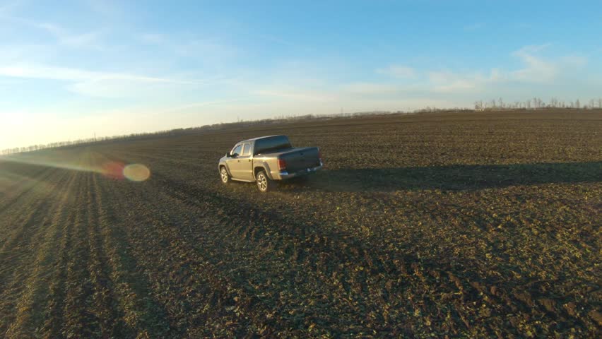 Off road vehicle riding along ploughed meadow. Pickup truck driving through plowed field at sunset time. Black car going on route with sunlight at background. Concept of agronomy farming. Aerial shot