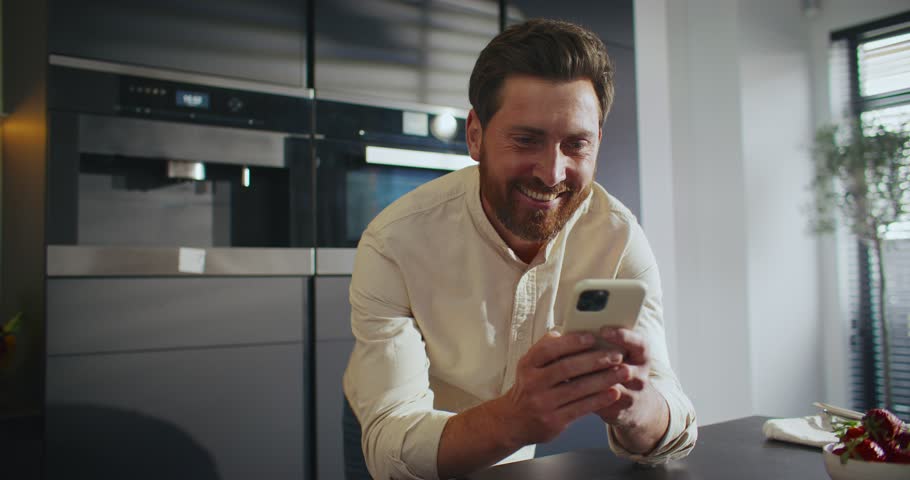 Zooming in on bearded Caucasian man intensively typing message on his cell phone. Enjoying online communication. Widely smiling after reading positive letter from friend or wife. Technology concept.
