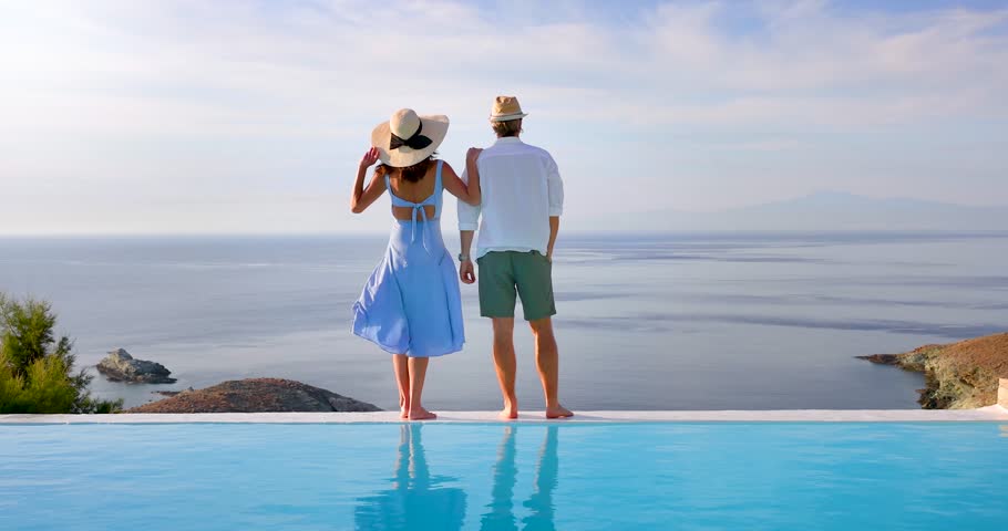 A romantic summer holiday couple stands at the edge of an infinity swimming pool and enjoys the view over the ocean