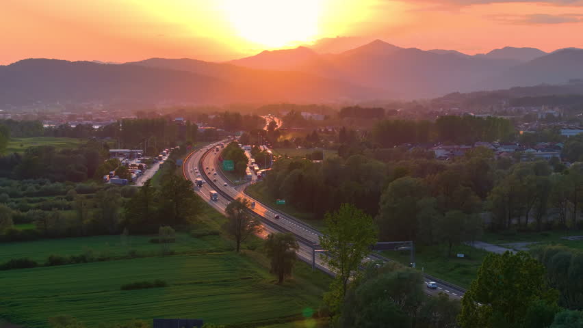 AERIAL, LENS FLARE: City highway passing through a scenic countryside at sunset. Ring road is lined with trees and distant mountains, with vehicles commuting in the warm evening light of setting sun.