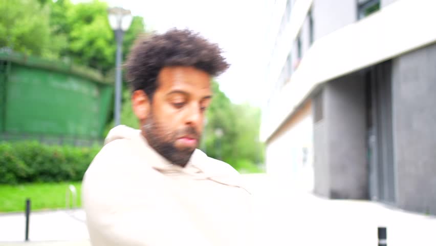 Black ethnic man dancing urban dance in the city with a beard and short hair is sitting on a bench. He is looking at his phone