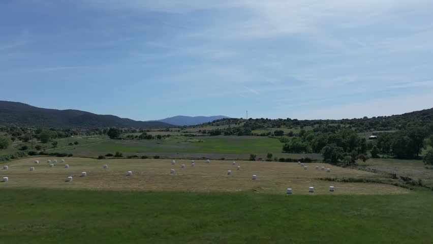 drone flight over a rural area with green meadows separated by walls and in one of them the grass has been mowed and there are large round bales of straw covered with white plastic in Avila