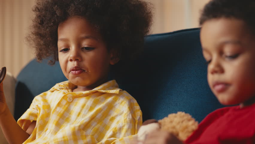 Two little African American boys eating cookies at home, enjoying sweet snack