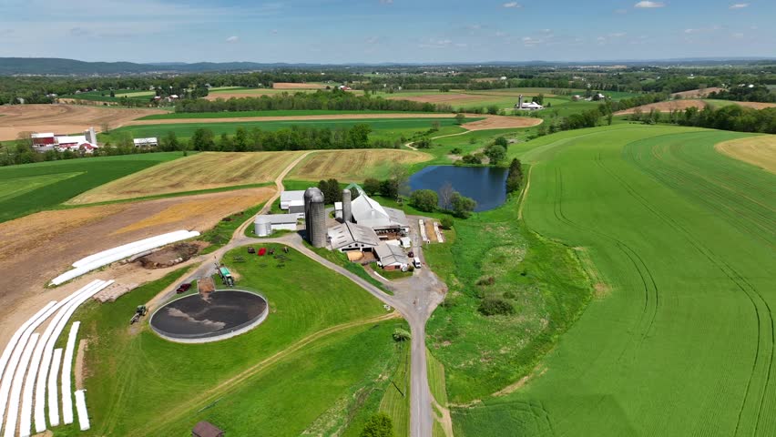 Aerial view of a serene countryside farm with silos, barns, a pond, and vast green fields, surrounded by rolling hills and distant farms. Shot in rural USA.