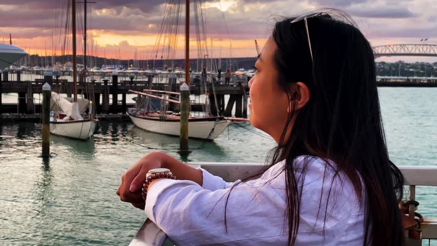 A smiling pretty Asian woman gazes peacefully at a sailboat harbor and water at sunset