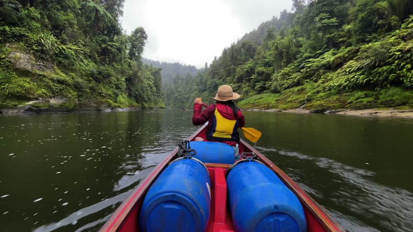 Woman in a red jacket, hat, and yellow life vest quickly paddles down the Whanganui River in her canoe between two forest cliffsides