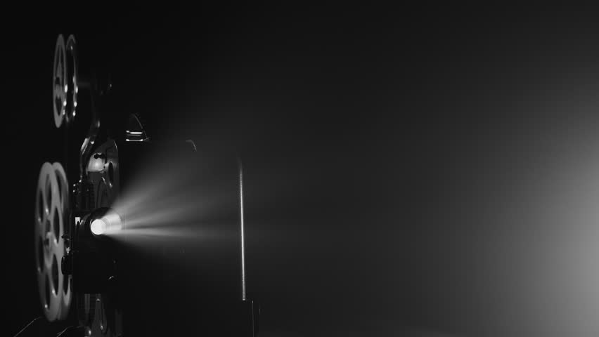 A black-and-white vintage movie projector working in the dark. A bright beam of light shines through the lens, illuminating the film reels. The atmosphere evokes classic film screenings