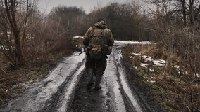A Ukrainian soldier walks down a muddy road in a forest area. He is dressed in military uniform and carries a backpack and gear. There are remnants of melting snow on the ground - Powered by Shutterstock - Get 15% off with code: PIKWIZARD15
