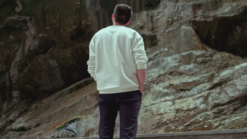 Man stands on background of rocky waterfall. Stock footage. Man at cliff with small waterfall. Man on trip in rocky canyon with waterfalls