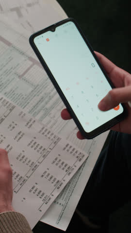 Close-up vertical shot of hands of young unrecognizable man holding bank account statement, pointing to numbers, adding costs on calculator on smartphone, while allocating personal budget
