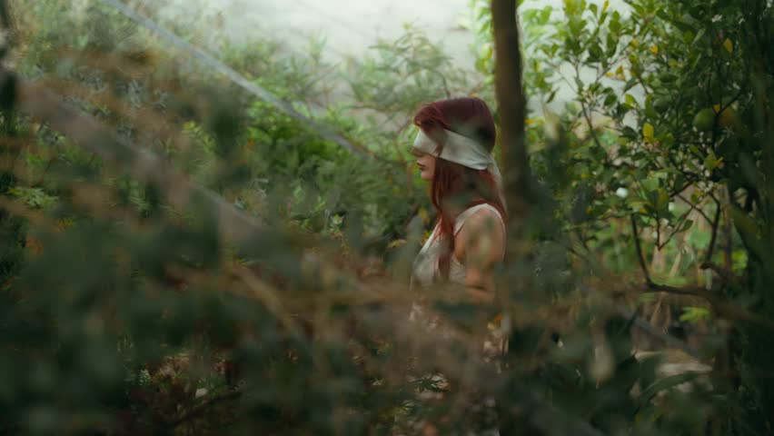 trucking cinematic footage with foreground focus of A young woman in a white dress and blindfolded while walking in a greenhouse of plants