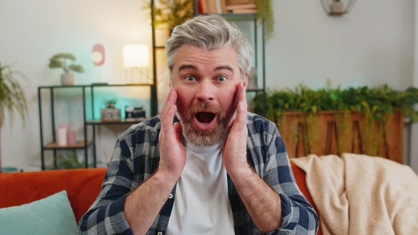 Wow emotion of bearded senior old man surprised looking at camera with big eyes, shocked by sudden victory, good win news, celebrating. Portrait of excited amazed mature guy sitting on sofa at home.