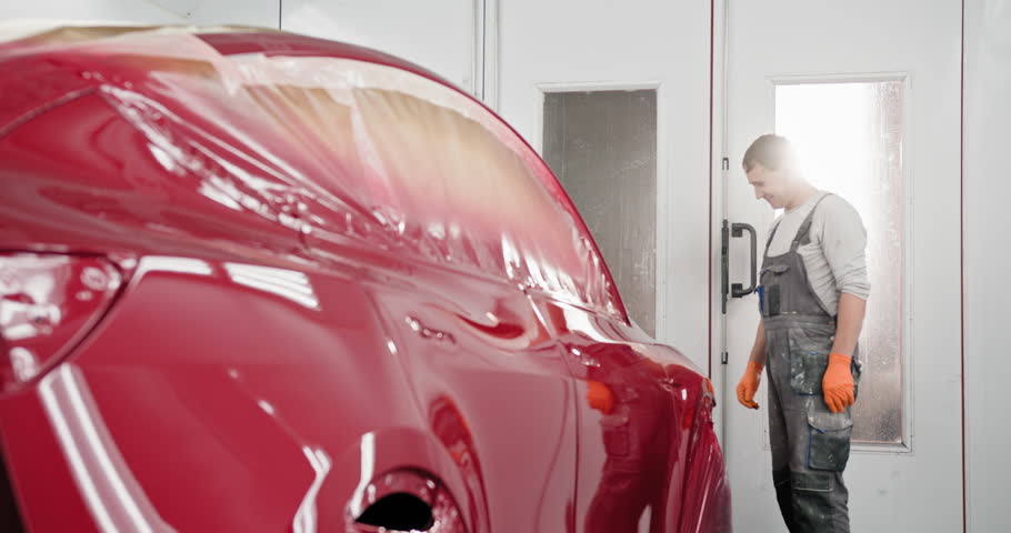 Automotive paint technician inspecting a newly painted red car in a specialized spray booth. Quality auto body shop concept showcasing professional car painting services. Technician Inspecting Car