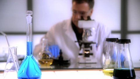 Focus remains on a volumetric flask and other lab glassware while a man working with a microscope works in the background.