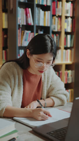 Asian girl reading information on laptop screen and writing notes in copybook as studying in college library. Vertical format clip