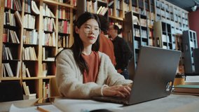 Time lapse of young Asian female student sitting at desk in public library and typing on laptop, people walking behind and taking books from shelves - Powered by Shutterstock - Get 15% off with code: PIKWIZARD15