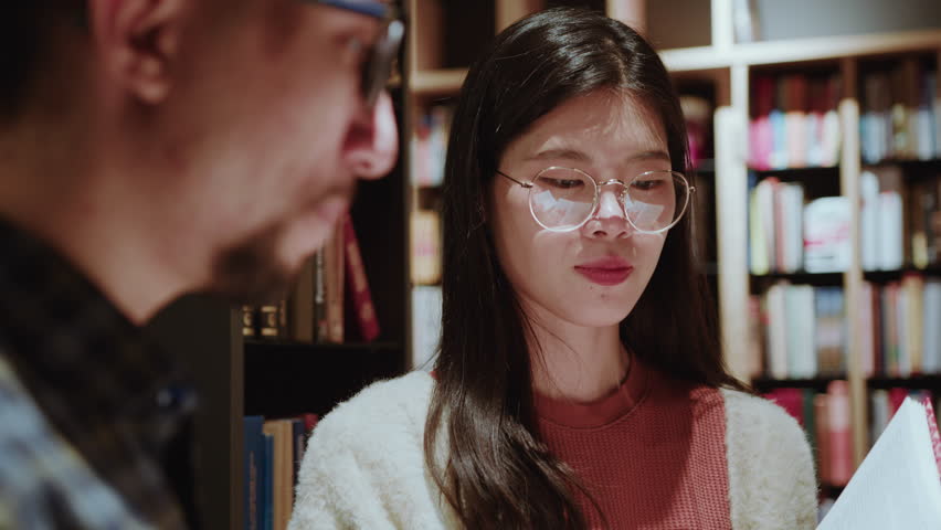 Young Asian female student standing in public library and discussing a book with male group mate. Over the shoulder view