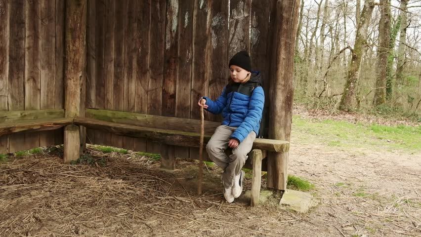 A young boy sits on a wooden bench in the woods, lost in thought as he rests his hand on a walking stick.