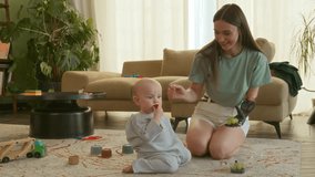 Young mother with prosthetic feeding her little son sitting on floor during playing toys in living room, she holds grapes in prosthetic hand and gives baby berries. Leisure together at home - Powered by Shutterstock - Get 15% off with code: PIKWIZARD15
