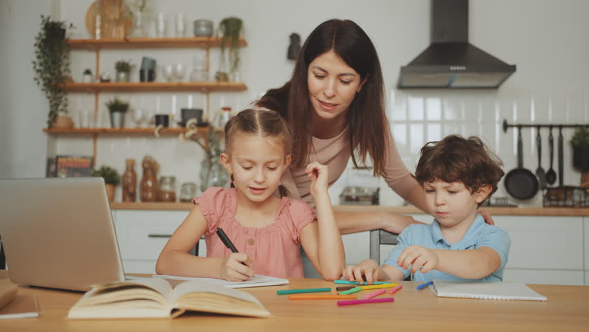 Mother assists two children with homework at table. One child writes in notebook while other plays with crayons. Concept of homeschooling and parental involvement. Learning and education at home - Powered by Shutterstock - Get 15% off with code: PIKWIZARD15