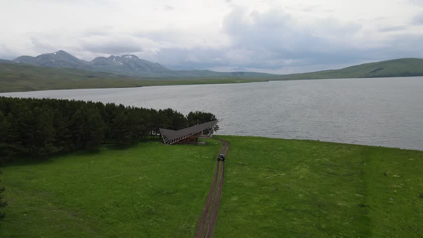 A view of a serene lake surrounded by lush green fields and distant mountains under a cloudy sky in Georgia. A small wooden structure extends towards the water, blending into the tranquil landscape.