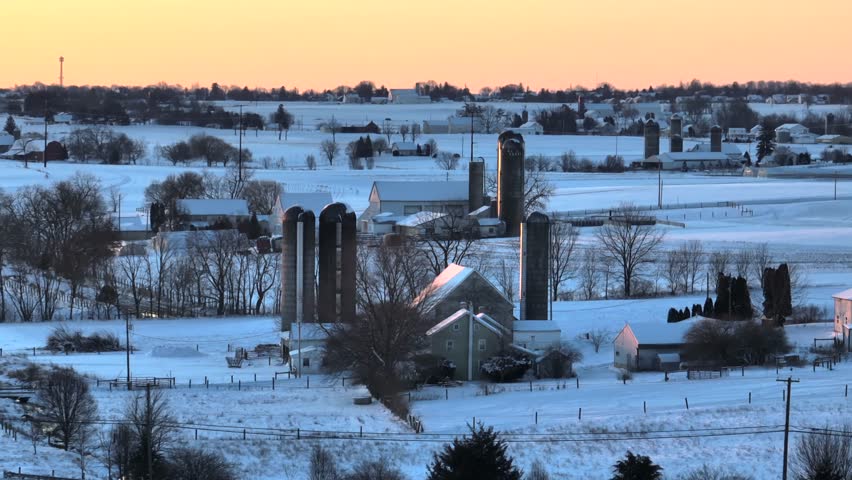 American countryside during winter sunrise with snow covered landscape. Silo and farm house in snowy winter landscape at morning. Aerial wide shot over rural country fields.