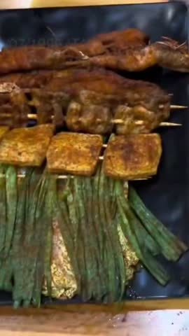 Close up of various Chinese street food skewers including grilled chives, tofu, and meat seasoned with spicy chili powder on a black platter.
