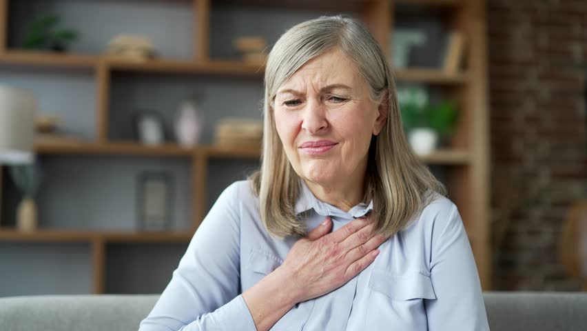 Senior female with gray hair suffering from heartburn sitting on sofa in living room at home. Sick elderly woman holding her hand on her chest and feels pain, hurts and burns in her stomach. Close up