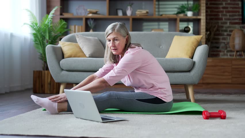Senior female practices gymnastics watching online course on laptop sitting on mat on floor in living room at home. Elderly woman doing stretching exercises listening to instructor via video call