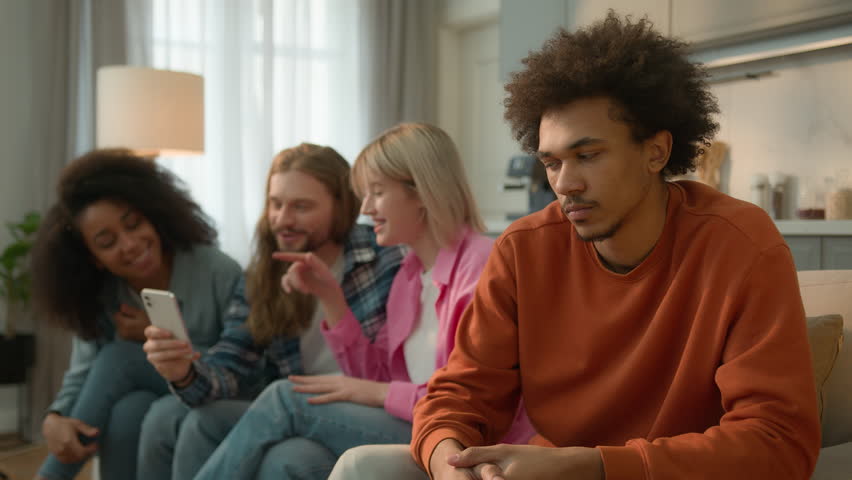 African American sad offended frustrated man guy male at home separated ignoring looking at group of multiracial Caucasian multiethnic friends women men mobile phone using addicted laughing gossiping