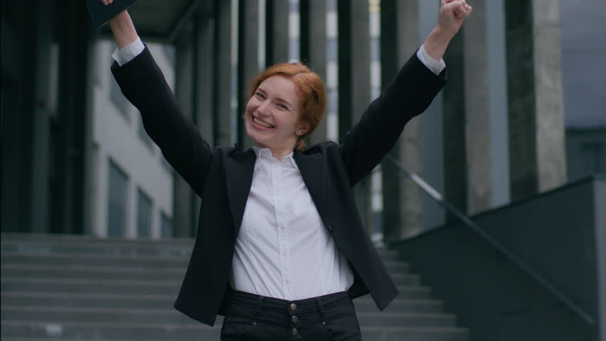 Overjoyed happy smiling young Caucasian girl businesswoman female student business woman winner raising arms hands celebrating promotion employment victory win gesture sincere emotion luck city street
