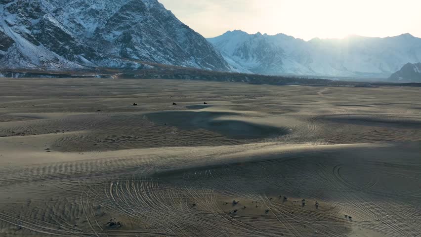 Aerial View of Sarfaranga Cold Desert landscape at sunset with sand dunes in the foreground and mountains with snow in the background and Vehicles Driving across. Push Forward Shot