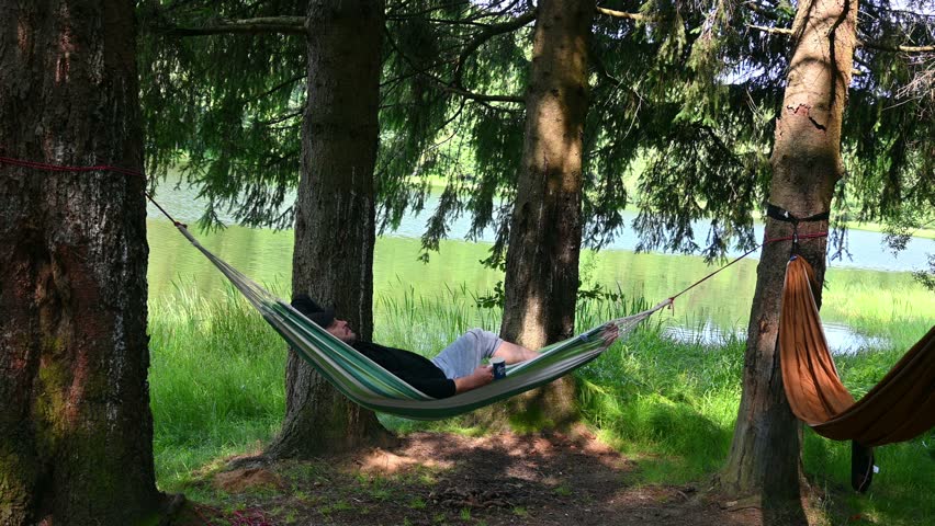 A man relaxes lying in a hammock, in a beautiful landscape, on the shore of the lake, among the trees. In camping, in nature. The man drinks tea. - Powered by Shutterstock - Get 15% off with code: PIKWIZARD15