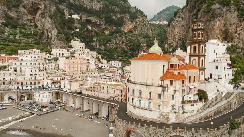 Historic buildings of Atrani perched on the cliff with winding road and arched bridge over sandy beach in summer day.