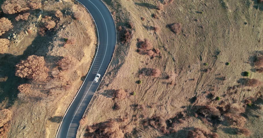 Winding mountain rural road with a white car driving down it at sunset, travel concept aerial shot