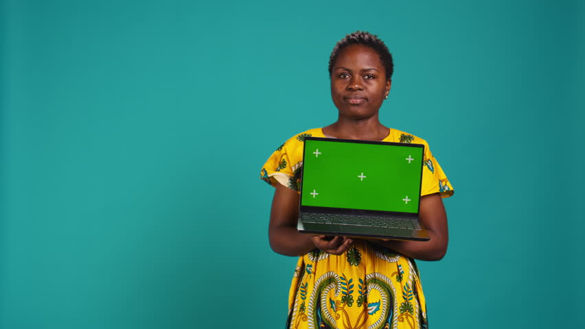 Confident smiling woman presenting a chroma key display on laptop, standing against blue background. Young adult holding a wireless pc and showing a green screen mockup. Camera B.