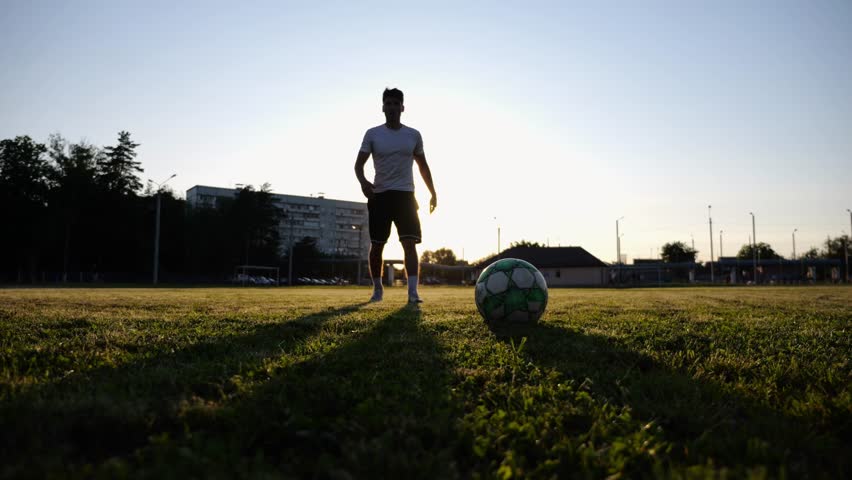 Silhouette of sportsman shooting a penalty kicks on stadium at sunset. Professional footballer kicking soccer ball at green field. Young man training at meadow. Concept of a freestyle football