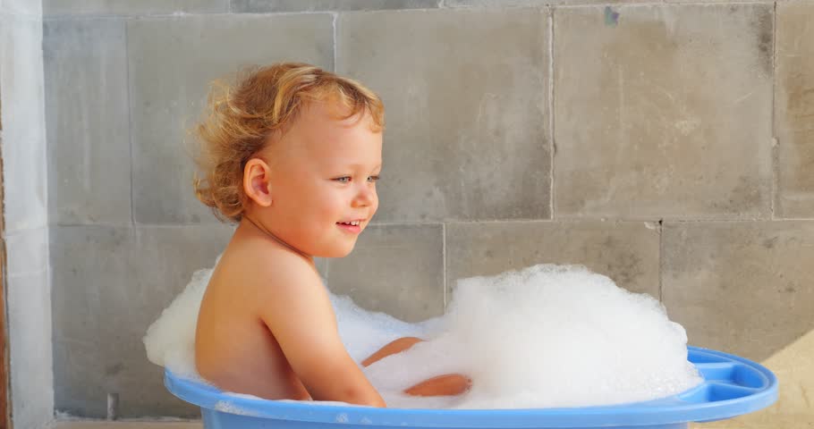 Smiling baby boy is resting in baby bathtub filled with warm water and white foam. Slow-motion footage captures delightful and peaceful moment of young child, portrayed from side.